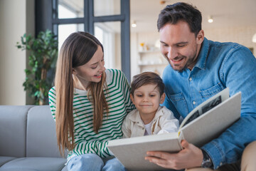 Friendly happy smiling family with little son boy kid, looking at family photo album together sitting in modern light living room at home. Emotional family moments, togetherness concept.