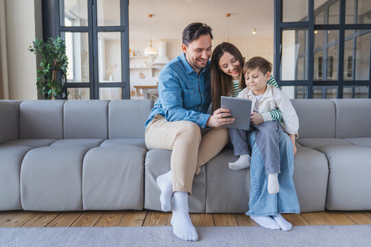 Portrait of Caucasian young family with child boy son sitting on the couch sofa at home using tablet. Smiling parents enjoying weekend together watching funny cartoon online or talking on video call. - Powered by Adobe