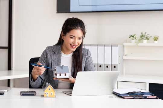 Online operation of buying and selling real estate. Young businesswoman realtor sits in the office and talks on a video call with clients, shows house model to the camera and offers product options.