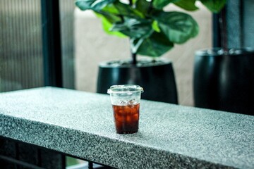 A clear plastic cup filled with iced coffee sits on a gray stone surface. Condensation forms on the outside of the cup, highlighting the cold beverage, with a blurred green natural background behind.