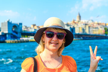 Young positive woman traveler in hat and sunglasses showing victory sign, smiling on city sea background. The concept of travel, adventure, freedom.