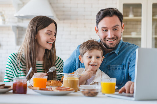 Family with little son sharing joyful morning meal around the kitchen table enjoying tasty food drinking orange juice watching video cartoon on laptop, eating breakfast together in modern apartment.
