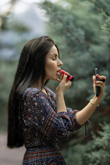 woman with long hair stands outdoors surrounded by greenery applying lip color as she takes a selfie. She wears a patterned dress and enjoys the moment in natural lighting.