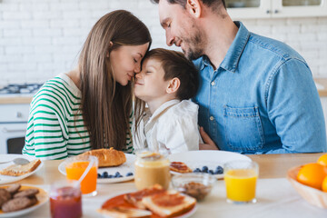 Happy family couple with child little boy having fun with son while sitting on table. Young smiling parents enjoying spending time with kid at the kitchen at home. Care love tenderness concept.
