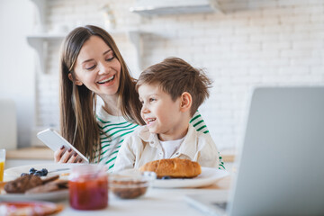 Family, gadget and breakfast kid with mother in a home happiness and care. Mom, food, laptop, smartphone and young child together with parent love and support with healthy drink and youth with a smile