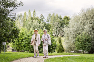 Two senior women friends walking together on park pathway, wearing light casual clothing and hats,...