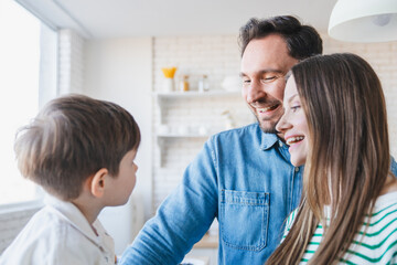 Fototapeta premium Joyful young parents and sweet kid boy standing in light cozy home interior, looking for son, laughing, smiling, promoting mortgage, property buying. Happy family concept