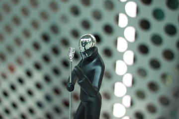 A sleek black figurine of a person holding a microphone stands against a metallic perforated...