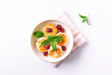 Cottage cheese pancakes with honey, raspberry and mint leaves on white ceramic plate on white marble table close up. Healthy homemade dessert.