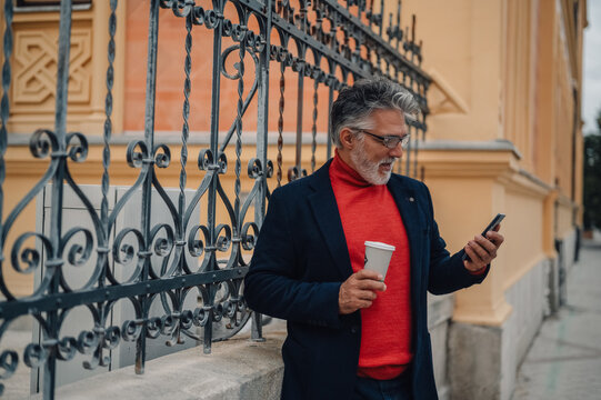 Stylish mature man using smartphone and drinking coffee outdoors