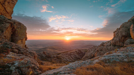Dramatic mountain landscape at sunset, viewed through a rocky gap, highlighting the sun setting over distant rolling hills and valleys under a brilliant sky with warm orange and cool blue tones