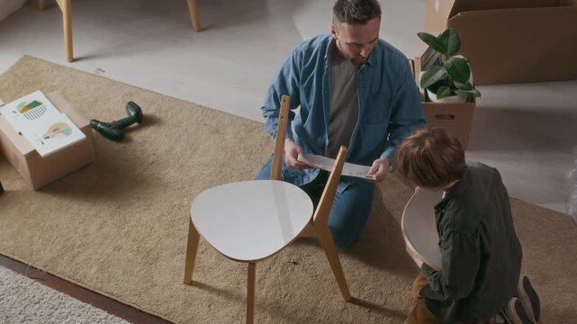 High angle shot of father and his son working together, reading instructions and assembling chair on floor