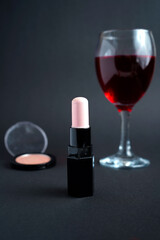 Light pink lipstick with black case placed beside compact powder and red wine glass, elegant beauty still life symbolizing femininity, glamour, and refined lifestyle.