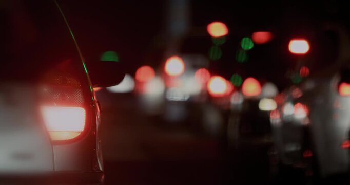 Rear of car with glowing red taillight, wide shot of night street with long blurred lines of traffic lights stretching into darkness, evoking urban delay