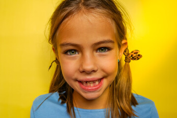 A smiling young girl with missing teeth, wearing a blue shirt, against a yellow background. She has brown hair and blue eyes. Baby teeth are being replaced by permanent teeth.
