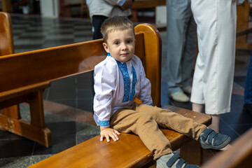 Little boy in traditional embroidered shirt sitting on church bench during christening ceremony....