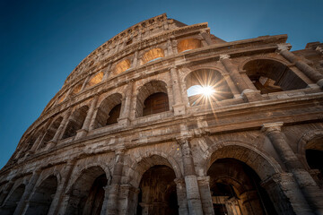 Fototapeta premium Ancient stone amphitheater with multiple arches under clear blue sky, sunlight shining through one of upper openings, showcasing historic architecture and grandeur