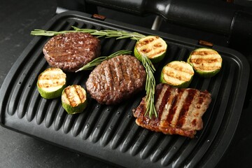 Electric grill with beef steaks, zucchini and rosemary on black table, closeup