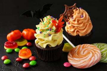 Delicious cupcakes with bat topper, sprinkles, chocolate web and colorful candies on dark textured table, closeup. Halloween celebration