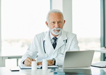 Obraz premium Portrait of a happy elderly male senior doctor using a laptop in his office in a hospital