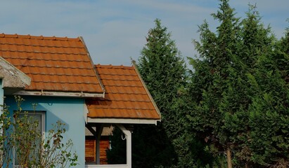 A small house with a red tiled roof next to tall evergreen trees under a blue sky.