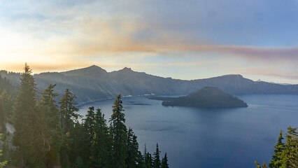 Aerial view of crater lake with wizard island and surrounding mountains under a hazy sky at sunrise