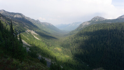 Obraz premium Aerial view of a mountain valley covered with dense green forests and rocky mountain peaks afar