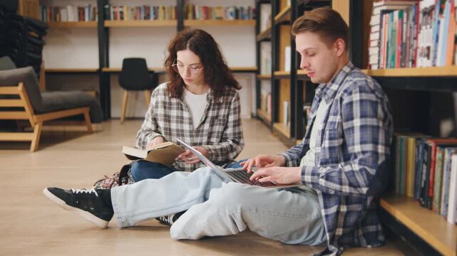 Two students studying together in a cozy library corner while seated on the floor surrounded by books on shelves
