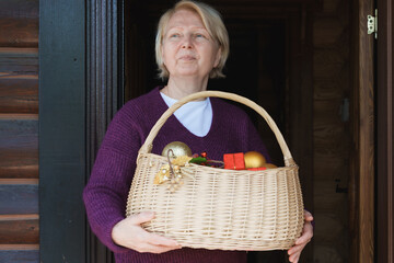 Elderly caucasian woman standing in doorway and holding a wicker basket filled with Christmas ornaments and festive decorations at home.