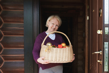 Elderly caucasian woman standing in doorway and holding a wicker basket filled with Christmas ornaments and festive decorations at home.
