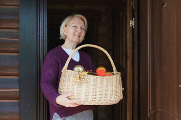 Elderly caucasian woman standing in doorway and holding a wicker basket filled with Christmas ornaments and festive decorations at home.