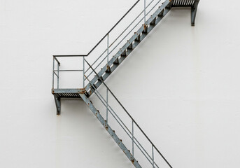 Durable industrial metal fire escape stairs ascend against a stark white textured building facade, suggesting urban grit and architectural detail.