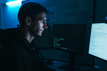 Caucasian male programmer in hoodie analyzing code on computer screens, working on cybersecurity and software development in dark office.