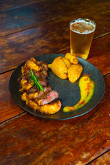 A mouthwatering steak, perfectly cooked and sliced, is served with golden roasted potatoes and a refreshing glass of beer, all on a rustic wooden table.