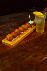 A wooden board holds golden-brown fried appetizers, served with a dipping sauce, alongside a glass of refreshing drink with an orange slice.