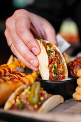 Close-up of a hand dipping a taco into sauce, surrounded by hot dogs with cheese and ketchup, and crispy fried snacks. Vibrant street food scene in casual dining setting