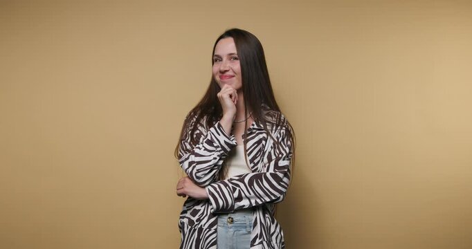 footage of young woman smiling while resting her chin on her hand, dressed stylish in a zebra print jacket on beige background
