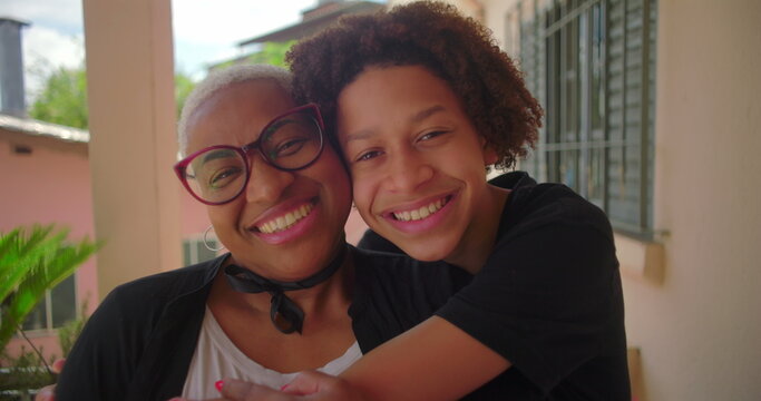 Warm embrace between an African American mother with glasses and her mixed-race curly-haired son on a porch in a warm, tropical neighborhood, joyful and affectionate family bond