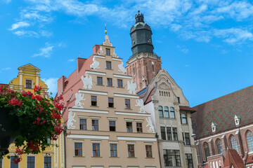 Fototapeta premium Ornate gabled buildings and St. Elizabeth’s Church dominate Wroclaw’s city center, with a lamppost and vibrant flowers in the foreground enhancing the picturesque urban landscape.