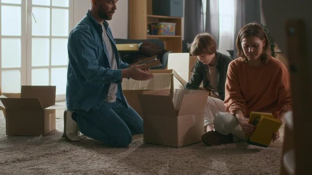 Family unpacking books from cardboard boxes and setting them up on shelves, settling into new apartment