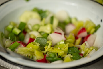 A close-up of a fresh vegetable salad served in a vintage white enamel bowl with a black rim. The salad includes sliced radishes, onion, cucumber, green onions, and bell peppers.