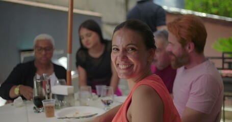 Woman turning her head towards the camera with a bright smile during a family BBQ lunch, seated outdoors with friends and family, enjoying a relaxed and cheerful gathering