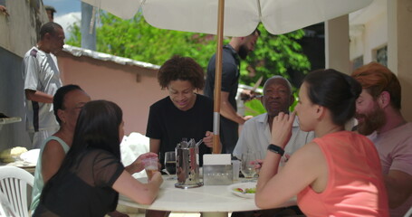 Multigenerational family enjoying outdoor meal, laughing and bonding at table under patio umbrella,...