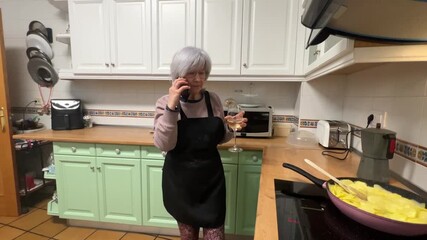 Senior woman with white hair enjoying a glass of wine and talking on phone while frying potatoes for Spanish omelette