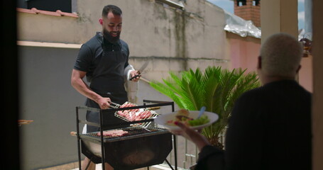 Man grilling meat on barbecue while Latin Hispanic woman of African descent eats salad, casual family meal outdoors in backyard with greenery