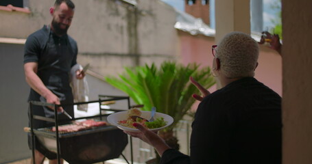 Man grilling meat on barbecue while Latin Hispanic woman of African descent eats salad, casual...
