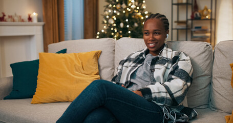 Fototapeta premium Attractive girl of African appearance wearing checkered shirt sitting on sofa in living room. Behind sofa shimmers Christmas tree. The girl listens to music in headphones and sings along.