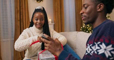 Sitting on a sofa, two friends of multi-racial background wear warm sweaters. The young woman wraps a gift with a red ribbon. The young man takes a photo, shows her the picture, and they smile and