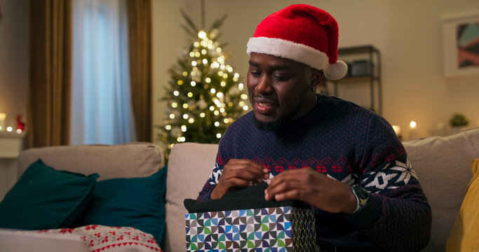 A smiling young man has a video chat with his girlfriend. He unpacks a gift, opens the box, pulls out a black scarf, is pleasantly surprised, and thanks the young woman.