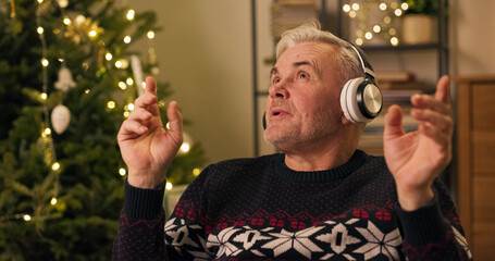 An attractive, happy middle-aged man with gray hair listens to Christmas carols. Dressed in a warm holiday sweater, he gestures with his hands, looks around, and sings as a shimmering Christmas tree
